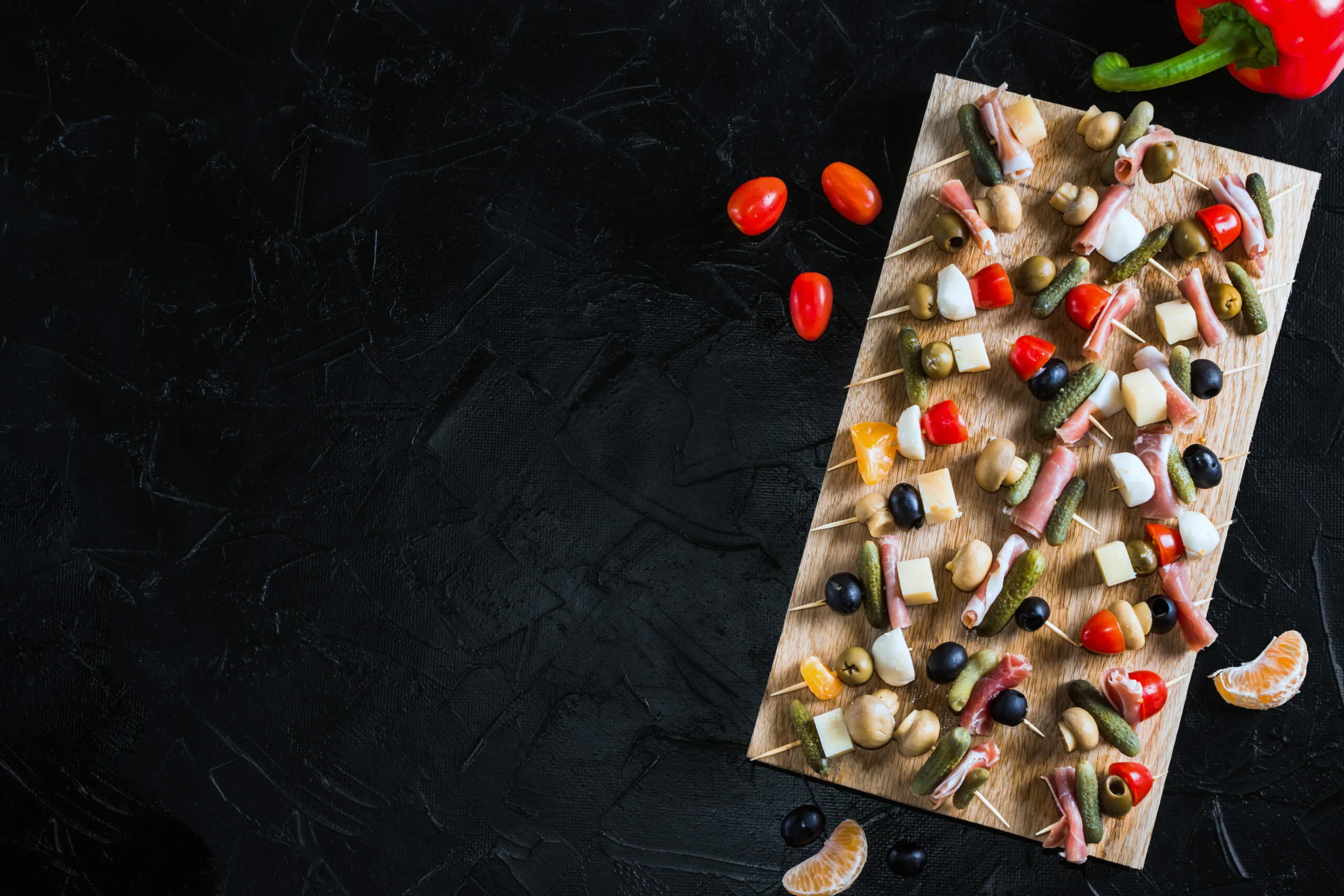 Snack skewers on a wooden tray on a black concrete table. Appetizers with mozzarella, ham, olives, cucumbers, tomatoes. Snack skewers on a wooden tray on a black concrete table. Appetizers with mozzarella, ham, olives, cucumbers, tomatoes.